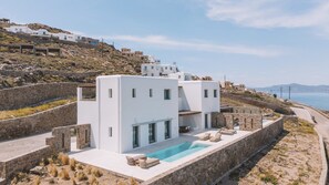 Eine moderne weiße Villa mit Swimmingpool, Steinmauern und Blick auf das Meer und die Hügel.