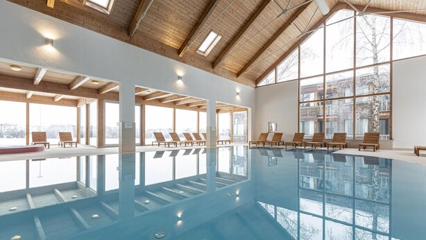 Una piscina interior con tumbonas de madera, ventanas amplias y un techo alto.