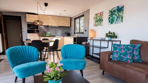 A modern living room with two blue armchairs, a brown sofa, and a coffee table with a plant. A kitchen with wooden cabinets and a dining area with a table and chairs is visible in the background. - Luxury Forest Lodge with Private Garden (Ravels)