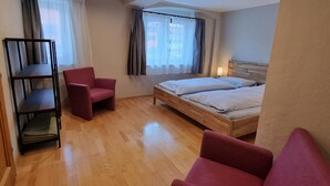 Hotellihuone, jossa on puulattia, valkoisilla lakanoiden peittämä sänky, punainen nojatuoli, pieni puupöytä ja kirjahylly.
