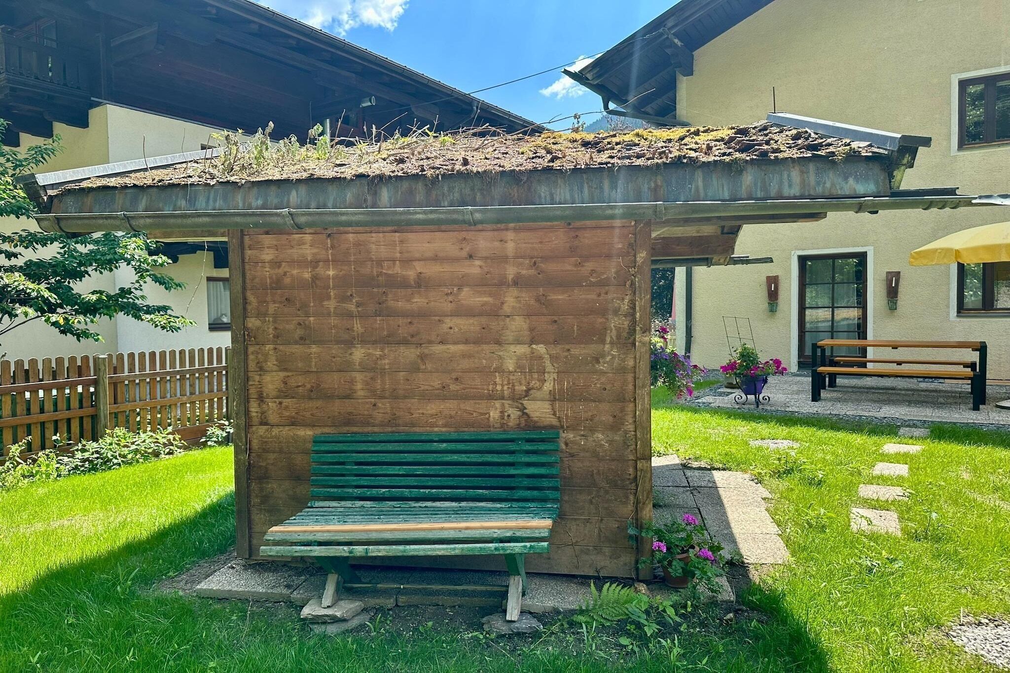 En träbänk under ett skydd med tak, ett trästaket och en byggnad med fönster och en dörr.