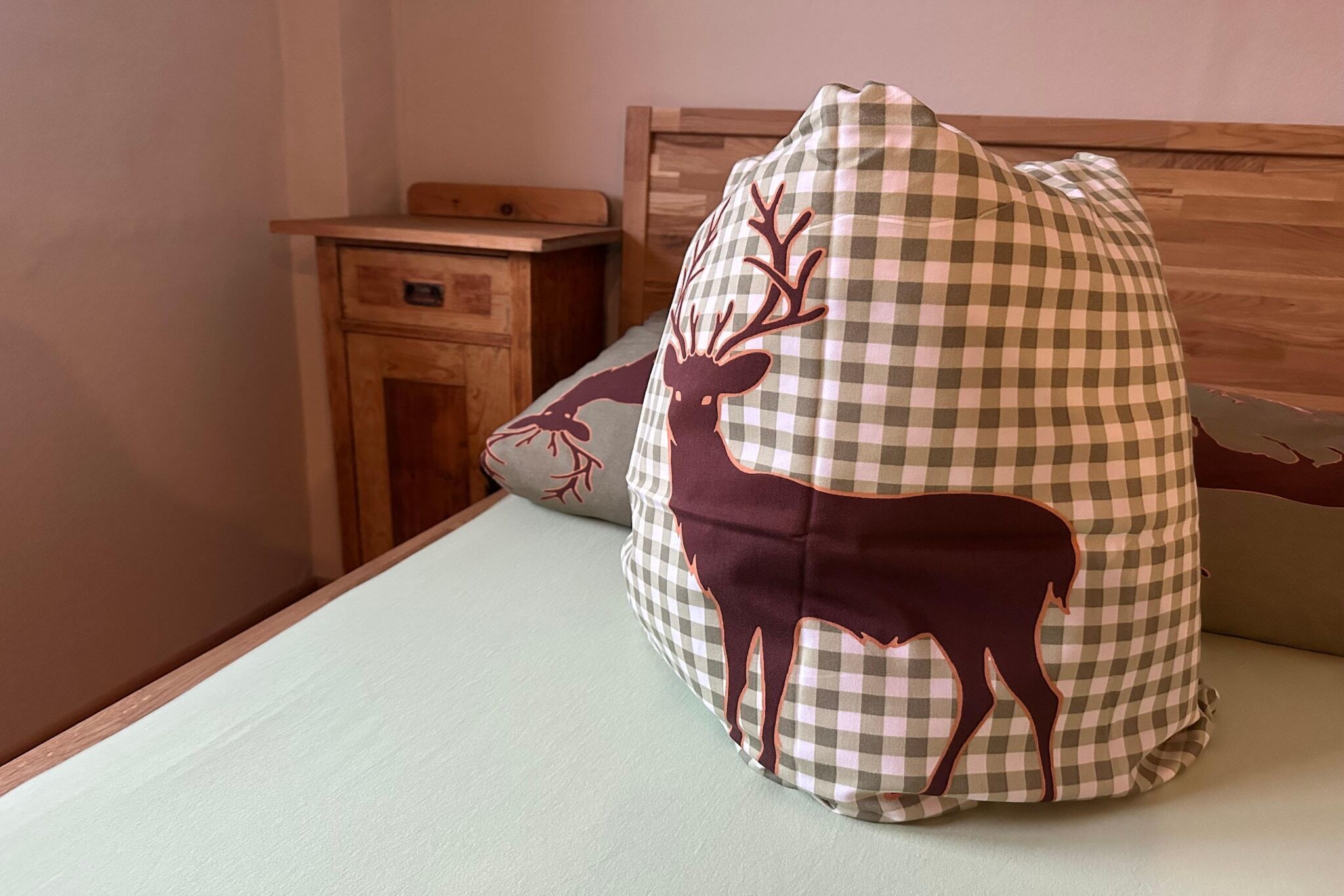 A bed with a checkered pillow featuring a deer design, a wooden nightstand with a drawer, and a plain wall.