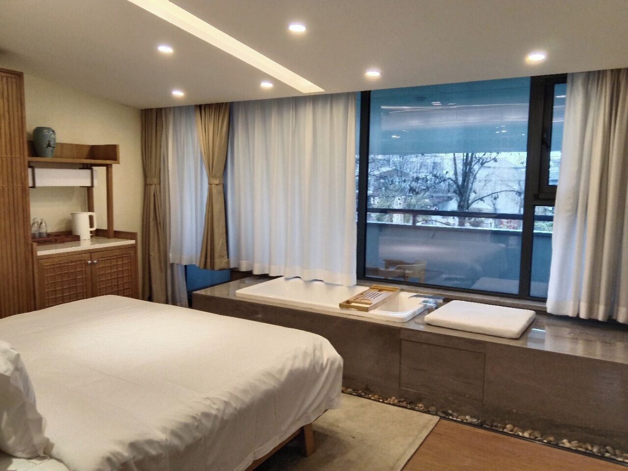 A hotel room with a large bed, a window with curtains, a small table, and a built-in bathtub.