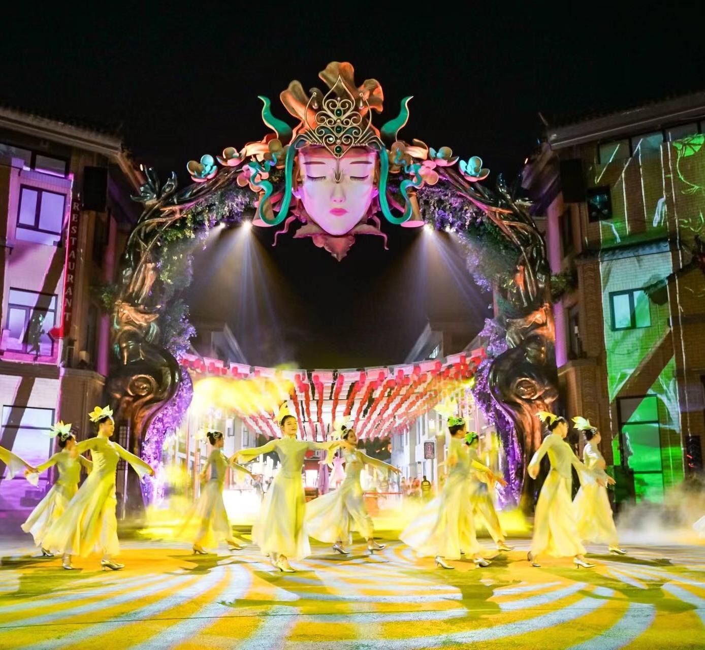 A traditional performance with dancers in yellow dresses, a large ornate mask, and a stage with a circular pattern.
