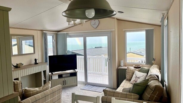 A living room with a television, sofa, and a view of the sea through a sliding door. - Waterside, Paignton - Holiday Accommodation 19143 (Paignton)