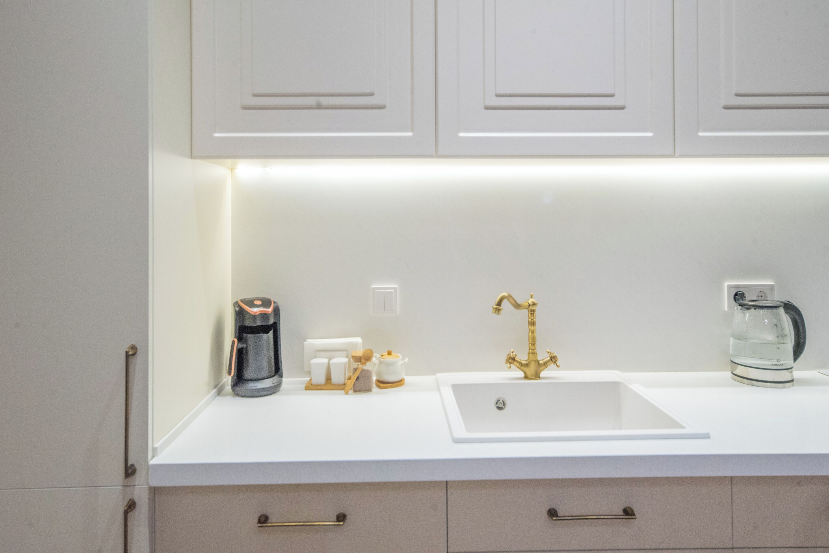 Una cocina moderna con armarios blancos, encimera blanca, grifo dorado, cafetera y hervidor.