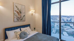 A bedroom with a bed, a nightstand, a wall-mounted lamp, a window with curtains, and a city view. - HB apartments University Agro (Tbilisi)