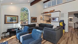 A spacious living room with a large window, a kitchen area, and a staircase leading to an upper level. - Steps to Snow Summit! LUX Tri-Level Condo! Ski Slope Views! Retro Arcade Games! (Big Bear Lake)