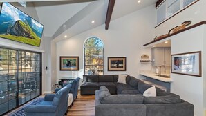 A spacious living area with a large sectional sofa, a small couch, and a coffee table. There is a large window with a view of a mountainous landscape. - Steps to Snow Summit! LUX Tri-Level Condo! Ski Slope Views! Retro Arcade Games! (Big Bear Lake)