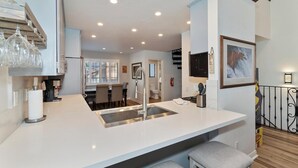 A modern kitchen with a central island, a dining area with a window, and a staircase leading to an upper level. - Steps to Snow Summit! LUX Tri-Level Condo! Ski Slope Views! Retro Arcade Games! (Big Bear Lake)
