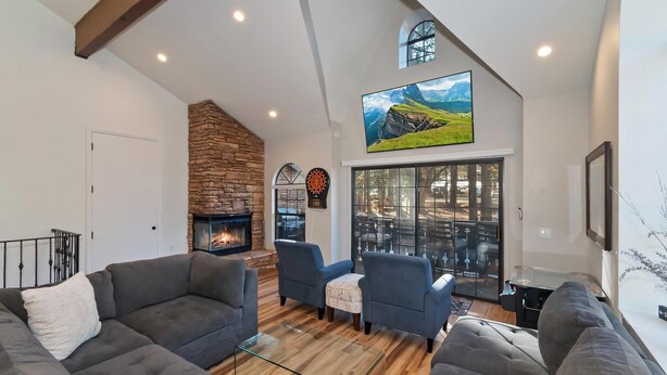 A spacious living room with a stone fireplace, comfortable seating, and a large window offering a view of the outdoors. - Steps to Snow Summit! LUX Tri-Level Condo! Ski Slope Views! Retro Arcade Games! (Big Bear Lake)