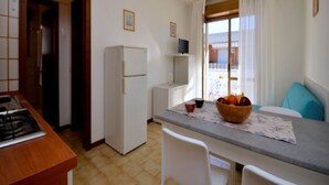Une cuisine compacte équipée d’une cuisinière à gaz, d’un réfrigérateur blanc, d’une table à manger avec un panier de fruits, et offrant une vue sur l’extérieur à travers une fenêtre ornée de rideaux.