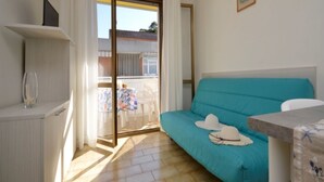 Une pièce avec un canapé de couleur turquoise, une petite table, un meuble blanc et une porte-fenêtre coulissante donnant sur un espace extérieur où se trouvent une chaise blanche et une table.