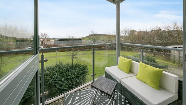 Un balcon doté d’une paroi vitrée, un canapé blanc avec des coussins verts, et une vue sur un espace verdoyant avec des arbres et une clôture.