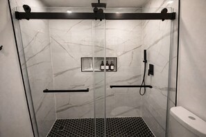 A modern shower enclosure with glass doors, a black towel rack, a handheld showerhead, and a small shelf with toiletries. - Sunny Oasis Near the Beach – 4BR/3BA Pool Home (Boynton Beach)