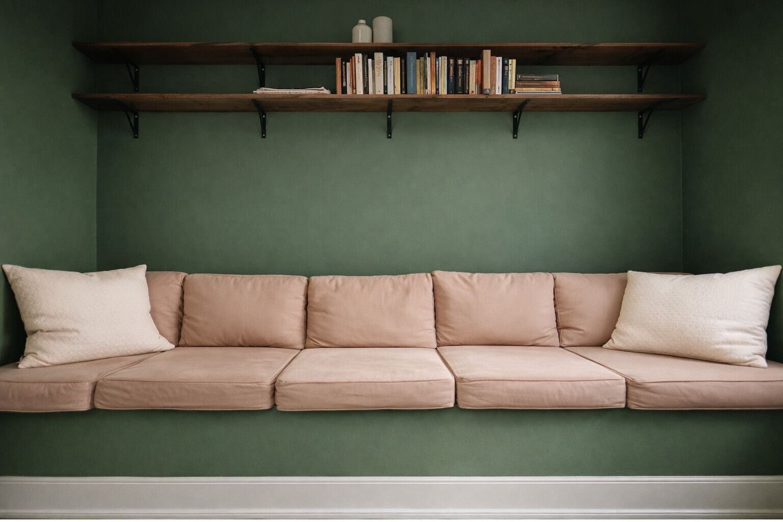 A beige sofa against a green wall with two wooden shelves above it, holding books and decorative items.