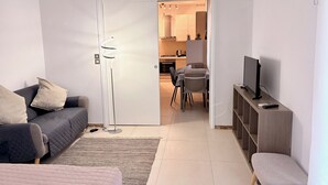 Ein modernes Wohnzimmer mit Sofa, einer Bank, einem Fernseher und einem Essbereich, der durch eine offene Tür sichtbar ist.