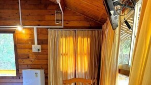 Interior de una cabaña de madera con una mesa de comedor, sillas, un refrigerador pequeño y cortinas.