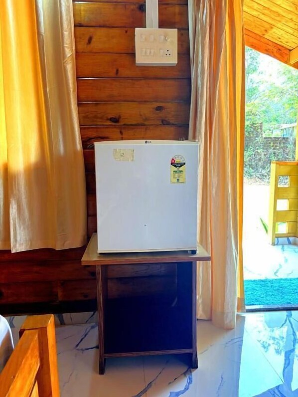 A compact room with a wooden wall, a small table with a white refrigerator, and a view of greenery outside. - Deer Cottages Agonda (Agonda)