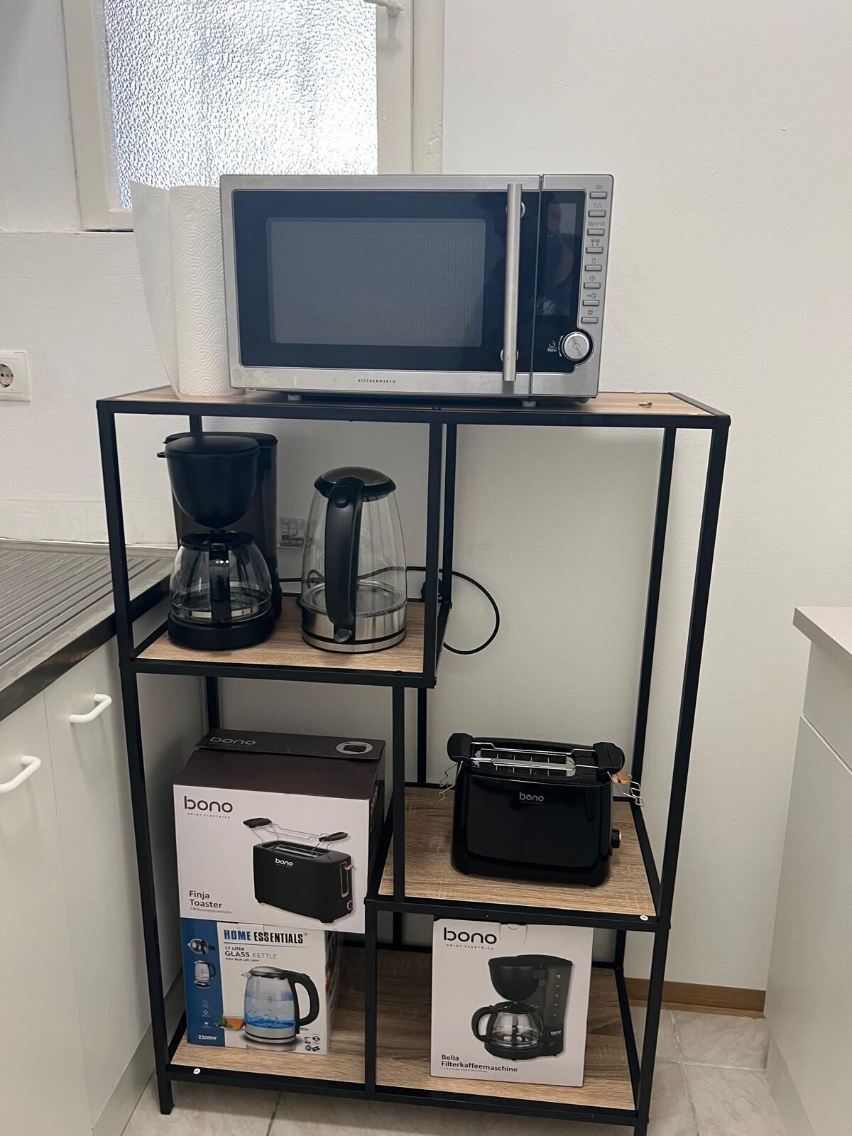 A kitchen setup with a microwave on top, a coffee maker, a toaster, and a kettle on a black metal shelf.