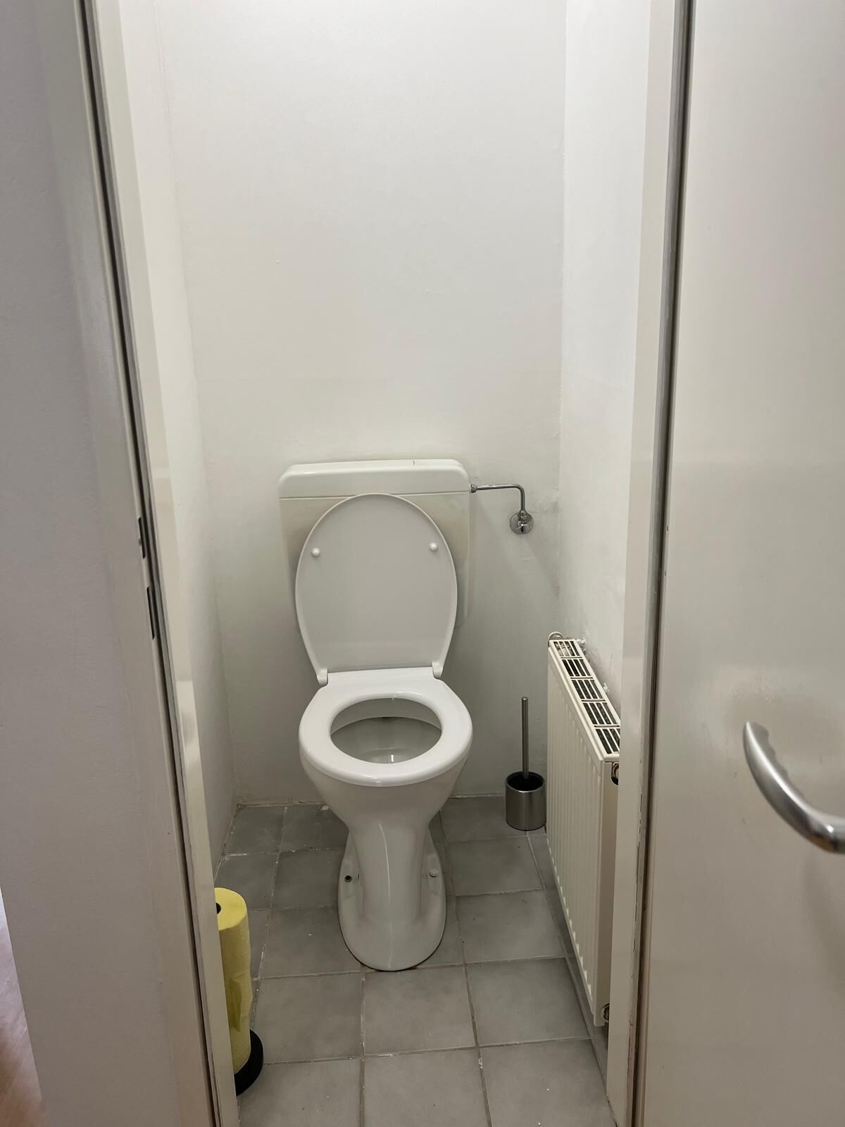 A small, white bathroom with a toilet, a toilet brush, and a yellow toilet paper roll.