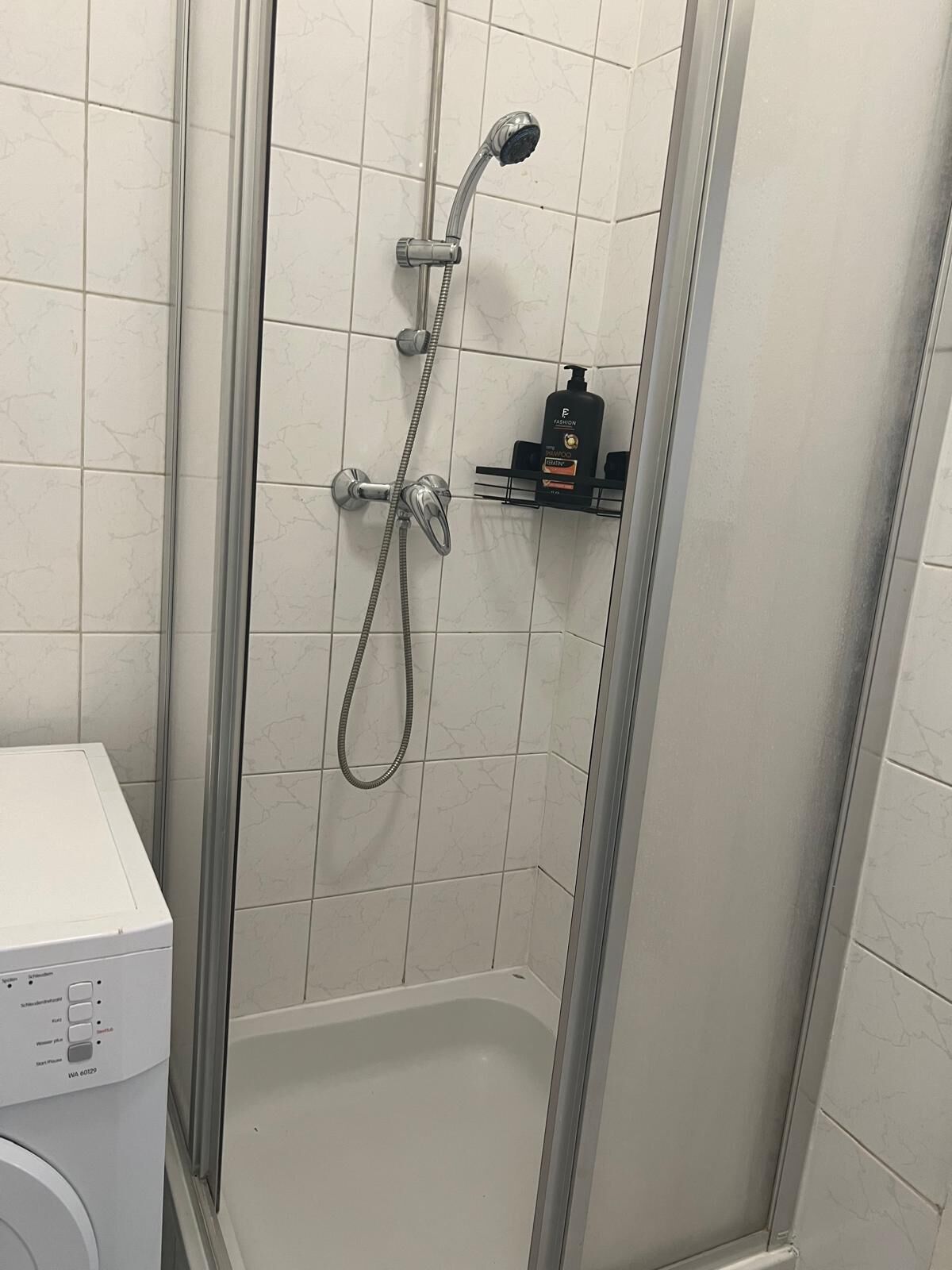Um chuveiro com chuveiro manual e torneira fixada na parede, parede com azulejos brancos e uma pequena prateleira com produtos de higiene.