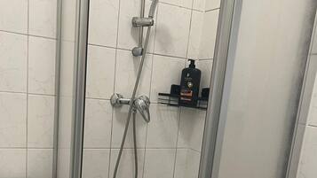 Um chuveiro com chuveiro manual e torneira fixada na parede, parede com azulejos brancos e uma pequena prateleira com produtos de higiene.
