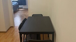 A room with a wooden floor, a dark-colored table with four chairs, a window with blue curtains, and a bed with a blue pillow in the background. - Modern 1BR City Apt for 5 Great Location (Vienna)