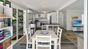 Uma cozinha moderna com armários brancos, um espaço de refeições com mesa de madeira e cadeiras brancas, e uma TV na sala de estar.