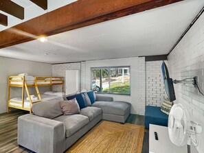 A modern room with a bunk bed, a grey sofa, a large window, and a white fan. - Family Beach House with Outdoor Entertaining (Long Beach)