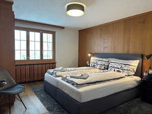 A bedroom with a large bed, a chair, a window, and a wooden wall. - Serene and relaxing 2 bedroom apartment with panoramic views (Saanen)