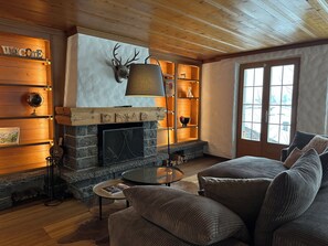 A cozy living room with a stone fireplace, wooden shelves, and a comfortable seating area. - Serene and relaxing 2 bedroom apartment with panoramic views (Saanen)