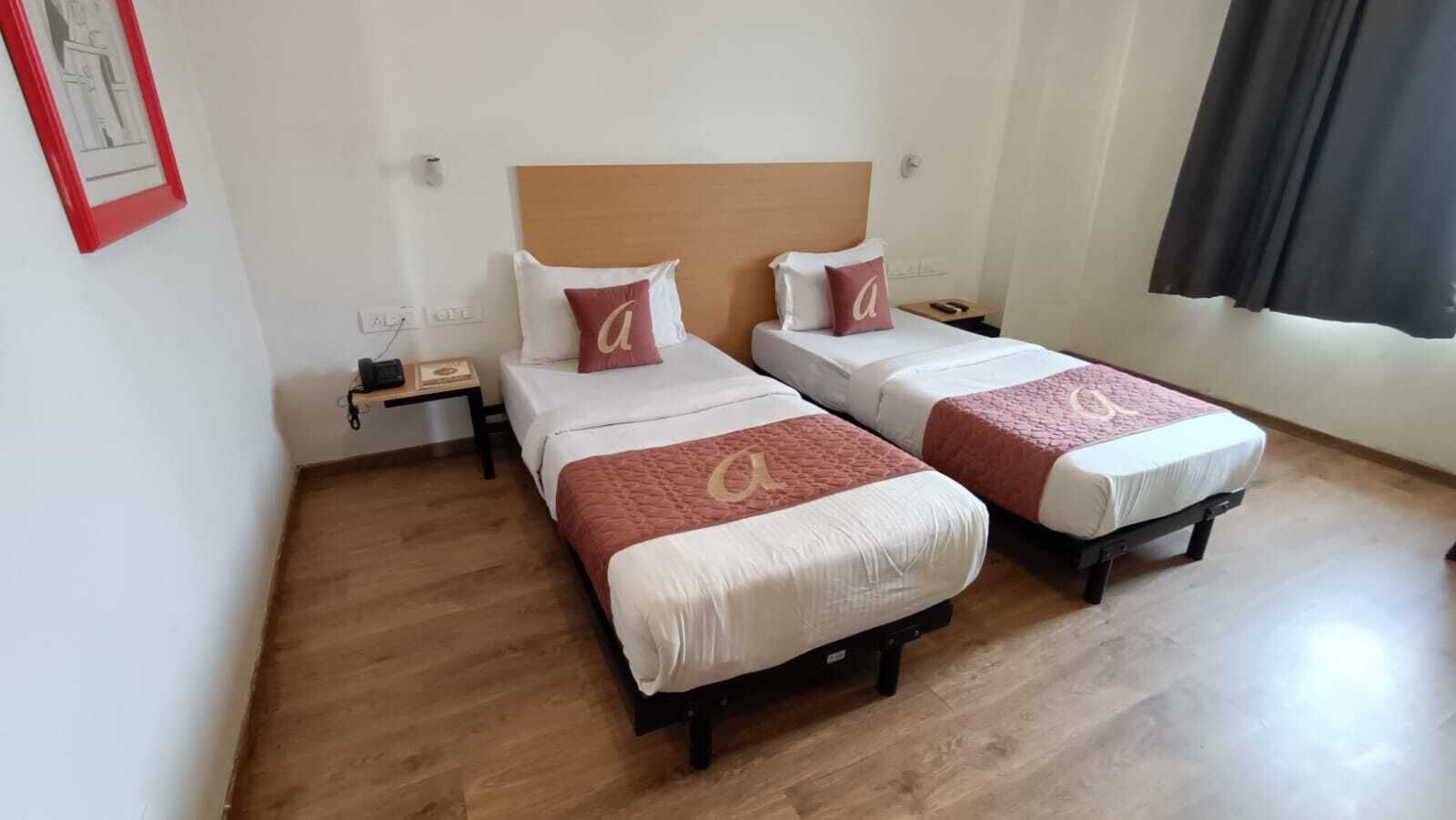 A hotel room with two beds, wooden flooring, a nightstand, and a framed picture on the wall.
