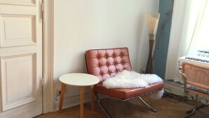 Ein Zimmer mit einem braunen, gesteppten Sessel, einem kleinen runden Tisch, einer weißen Tür und einem Fenster mit Vorhängen.