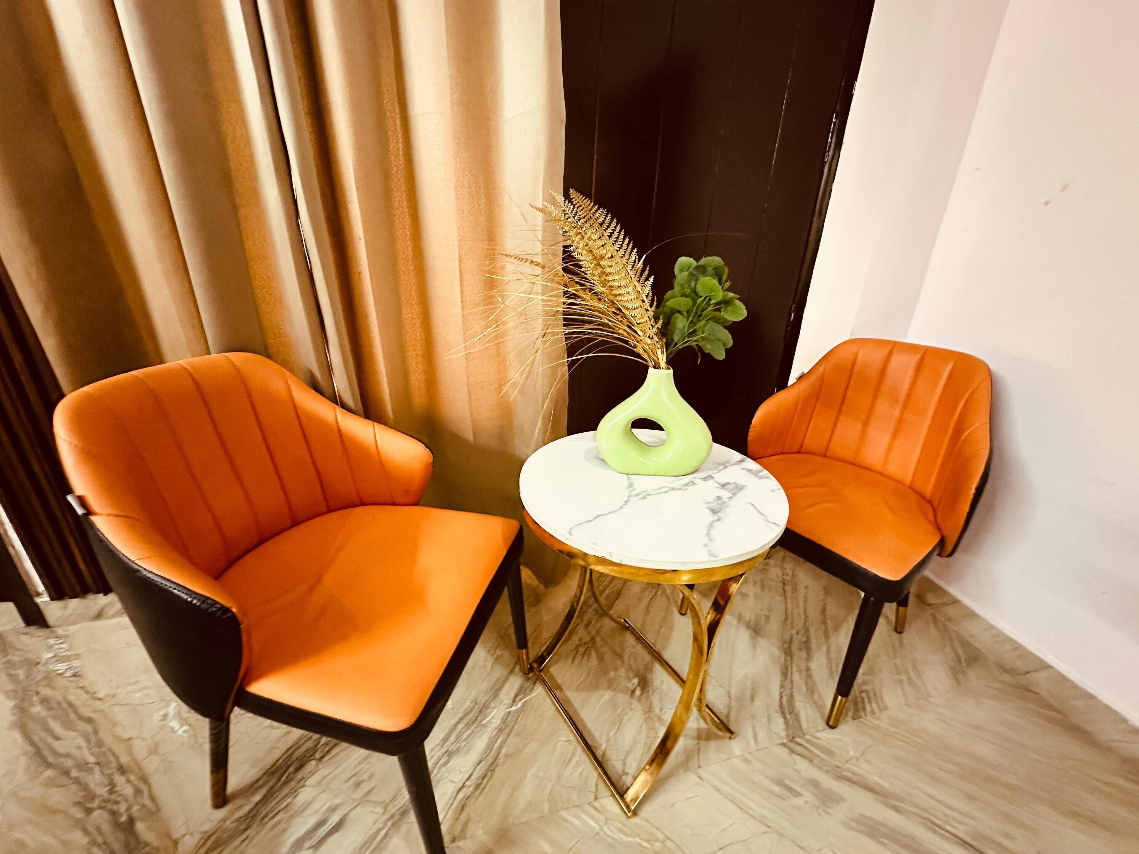 Deux chaises orange à armature noire et une petite table ronde ornée d’un vase vert et de plantes séchées.