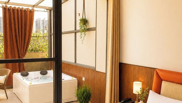 A hotel room with a bed, a small bedside table with a lamp, a nightstand with a phone, a window with curtains, a view of a cityscape, and a bathroom with a bathtub and a plant. - Savana Suites (Noida)