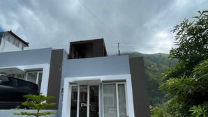 A modern two-story house with a paved driveway, a small garden with a tree, and a view of a hilly landscape.