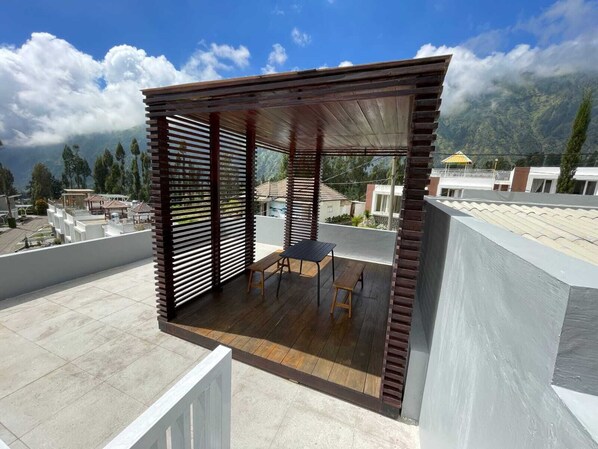 A modern outdoor patio with a wooden table and benches, overlooking a residential area with houses and mountains in the background. - Orlando Hill Villa (Ngadisari)