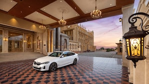 Une voiture de luxe blanche est garée sur un sol carrelé à motifs, devant un imposant bâtiment à l’architecture classique, et un lustre scintille au plafond.