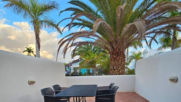 Une terrasse avec une table et des chaises, entourée de palmiers et d’un mur blanc.