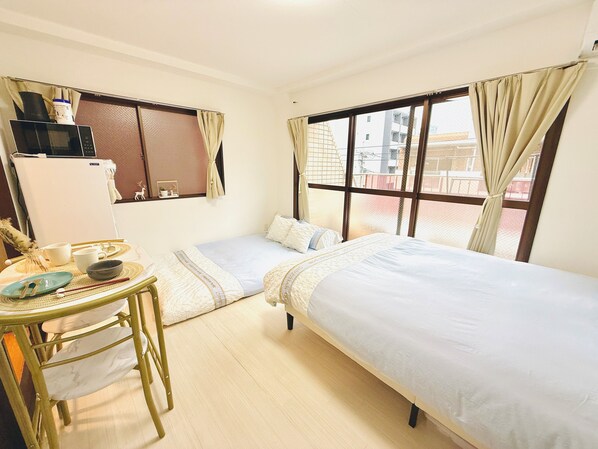Une petite chambre d’hôtel avec deux lits simples, une petite table avec une chaise, un four à micro-ondes et une fenêtre avec des rideaux.