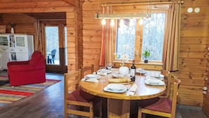 Ein Interieur einer Holzhütte mit einem Esstisch, gedeckt für vier Personen, umgeben von Holzsesseln. Im Hintergrund ist ein roter Sessel zu sehen, und ein Fenster mit Vorhängen lässt natürliches Licht herein.