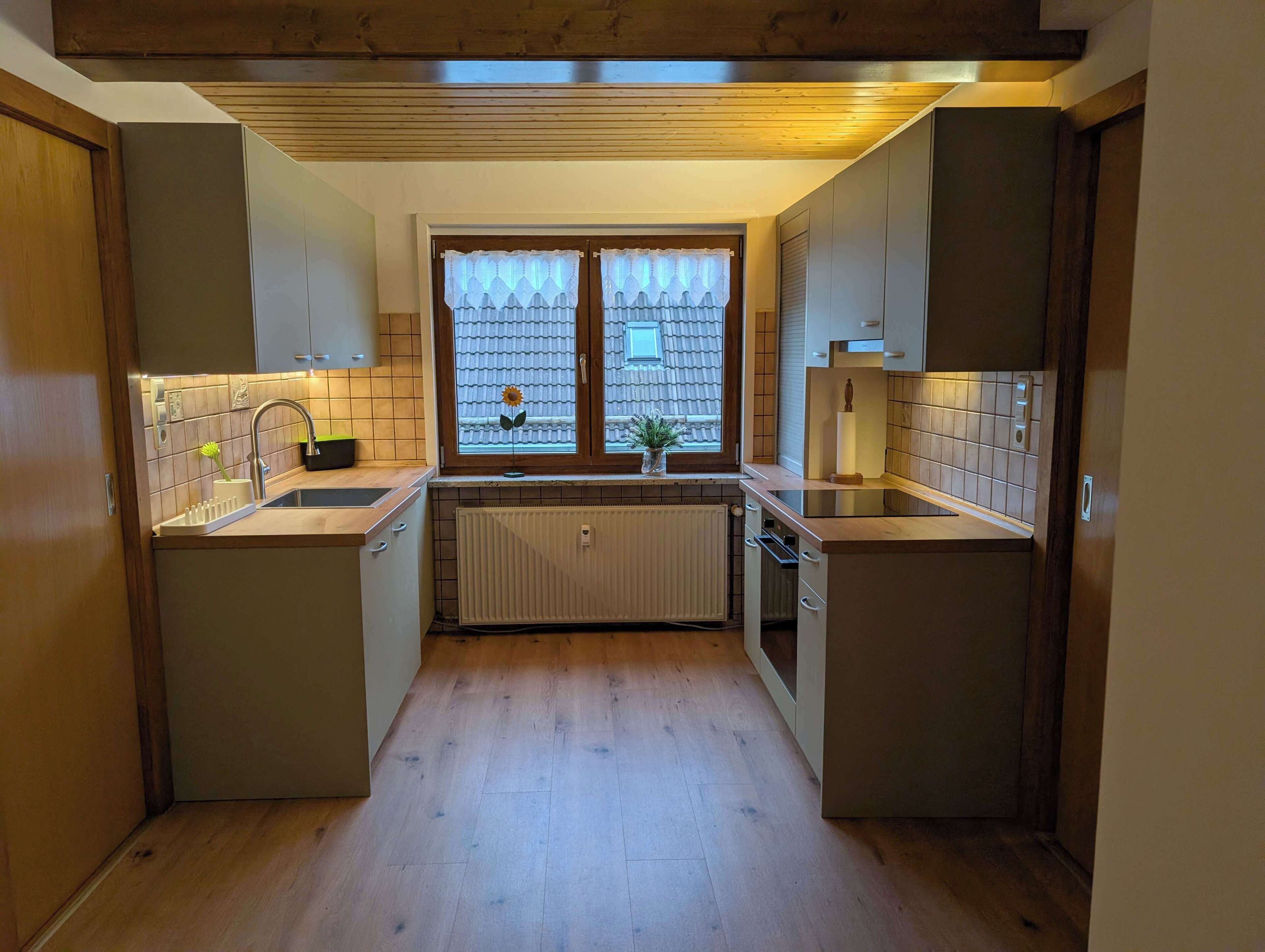 Eine kleine Küche mit Holzausstattung, einer zentralen Kochinsel, einem Fenster mit Vorhängen und einer Heizung.