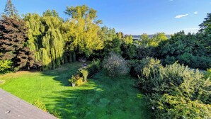 Ein gepflegter Garten mit verschiedenen Bäumen und Sträuchern, einem klaren blauen Himmel und Blick auf eine ferne Stadt.