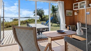 A spacious room with a large sliding glass door leading to a deck with a view of the water. Inside, there is a wooden table, a sofa, and a small cabinet. - "Rajna" - 30m from the sea by Interhome (Martofte)