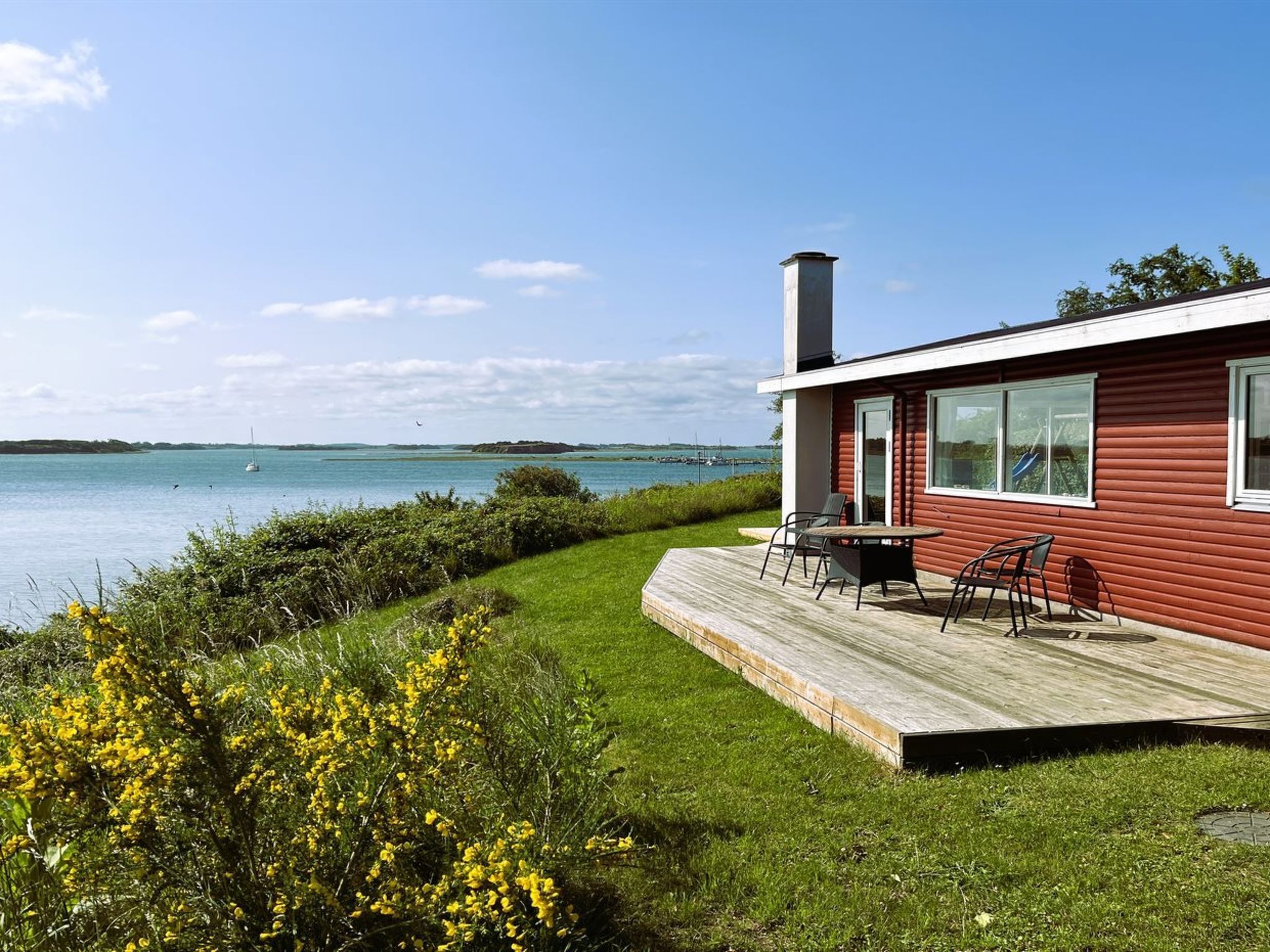 A red cabin with a deck overlooking a calm lake, surrounded by greenery and yellow flowers.