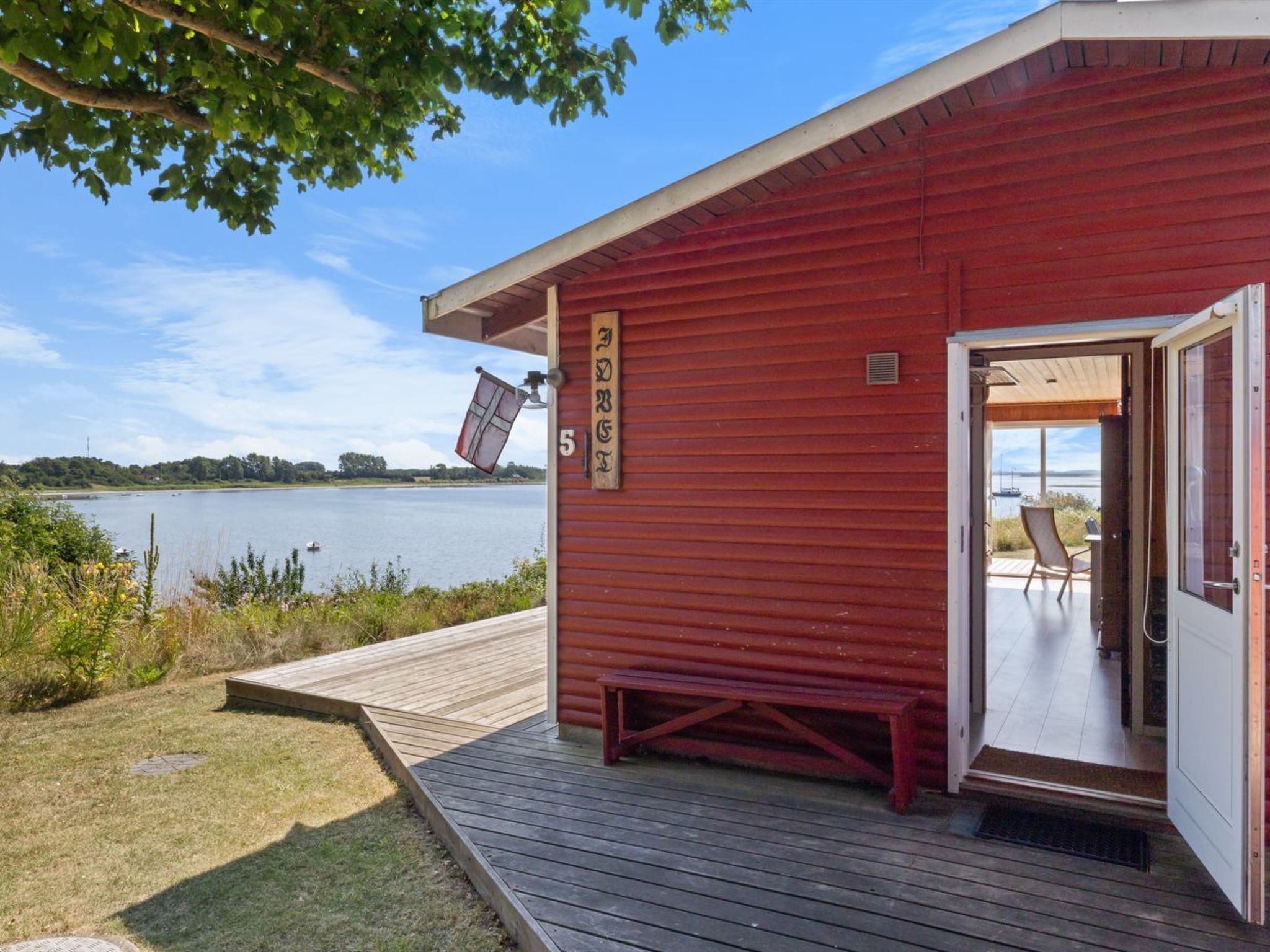 Una cabaña roja con una terraza de madera, un banco y una puerta que da a un interior con vista al lago.