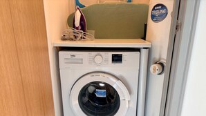 A compact, white washing machine with a control panel and a front-loading door, placed in a small laundry area with a green chair and an ironing board. - Bright 2-bed Apartment York City Centre - Walk to Everything (York)