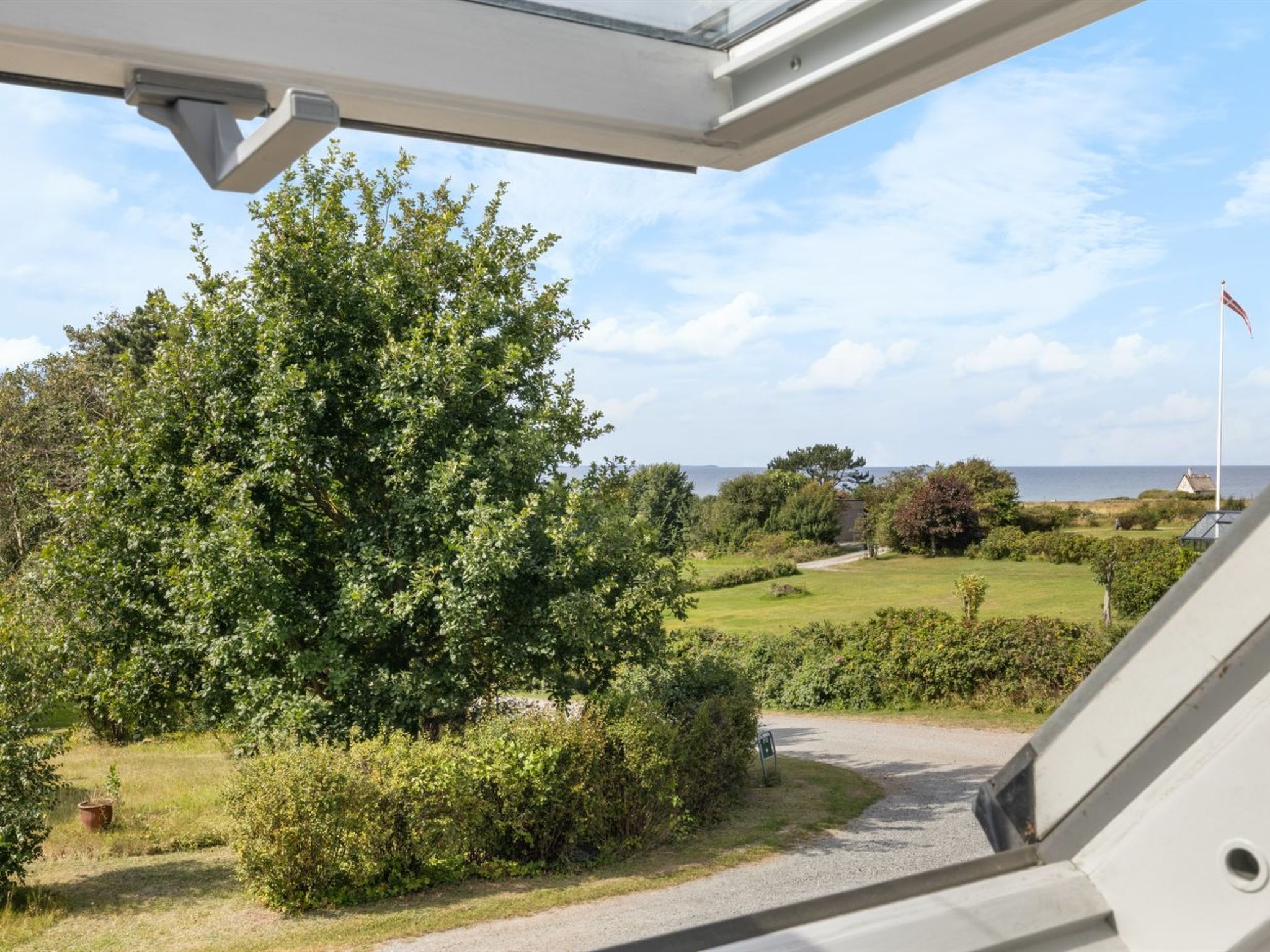 Une vue depuis une fenêtre donnant sur un jardin bien entretenu, avec une variété d’arbres et d’arbustes, un chemin de gravier et un mât sur lequel flotte un drapeau.