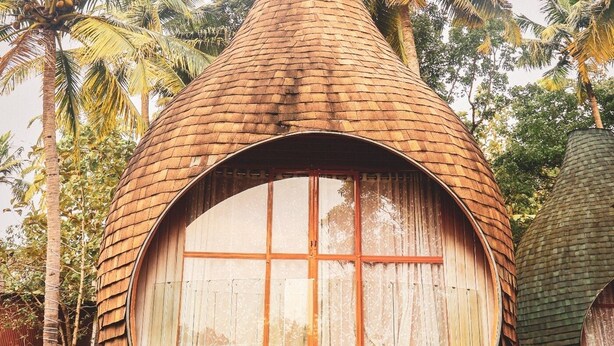 Une hutte au toit de chaume, dotée d’une grande fenêtre soutenue par des pieds en métal, et entourée de palmiers.